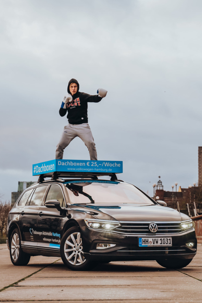 Auf einem VW ist ein Mann als Boxer mit Boxhandschuhen auf einer Fläche abgebildet, die normalerweise für eine Dachbox verwendet wird.
