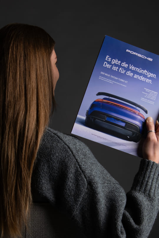 Eine Frau wurde von hinten fotografiert, während sie das Magazin 'Porsche Times' auf Schulterhöhe hält. Das Magazin zeigt die U4 mit einer prägnanten Headline und einem Porsche-Modell.