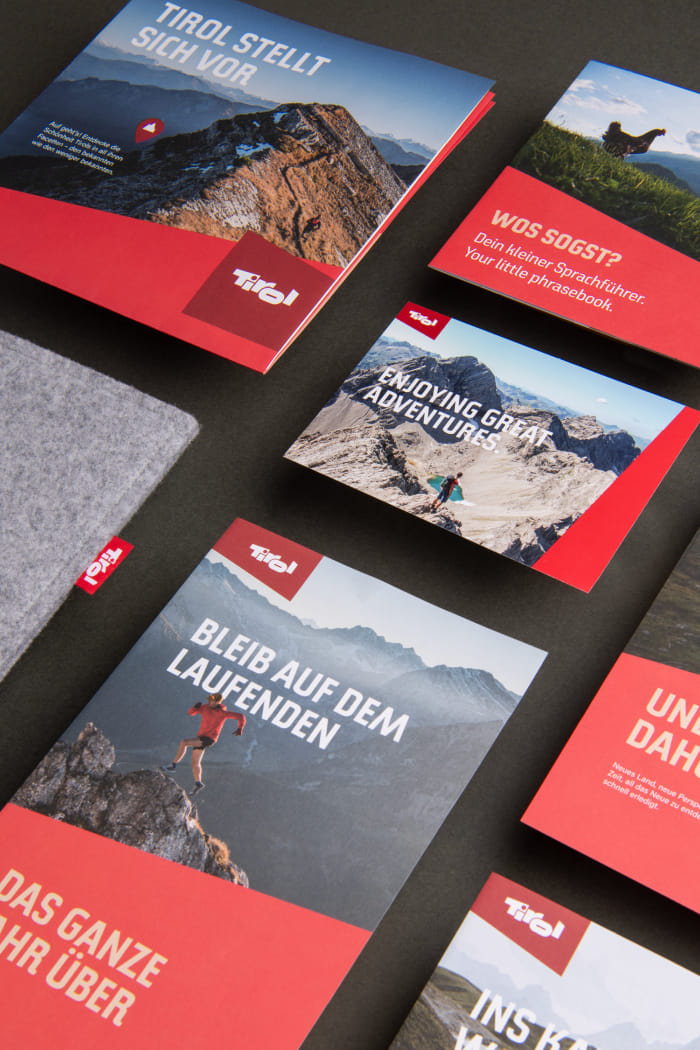 Mehrere Elemente aus dem Welcome Package sind auf einer Fläche aufgelegt. Darunter ist das Design der Vorderseite im Layout der Kampagne zu sehen, mit einer Fotografie eines Läufers am Berg, dem Logo sowie einer roten Fläche für den Titel.