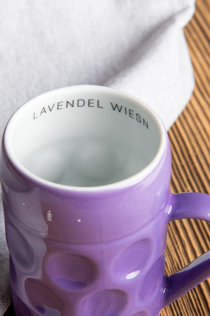 Ein kleiner Bierkrug in der Farbe der Provence (Lila) steht auf einem Tisch. Die Innenseite des Kruges ist weiß und mit dem Eventnamen 'Lavendel Wiesn' bedruckt.