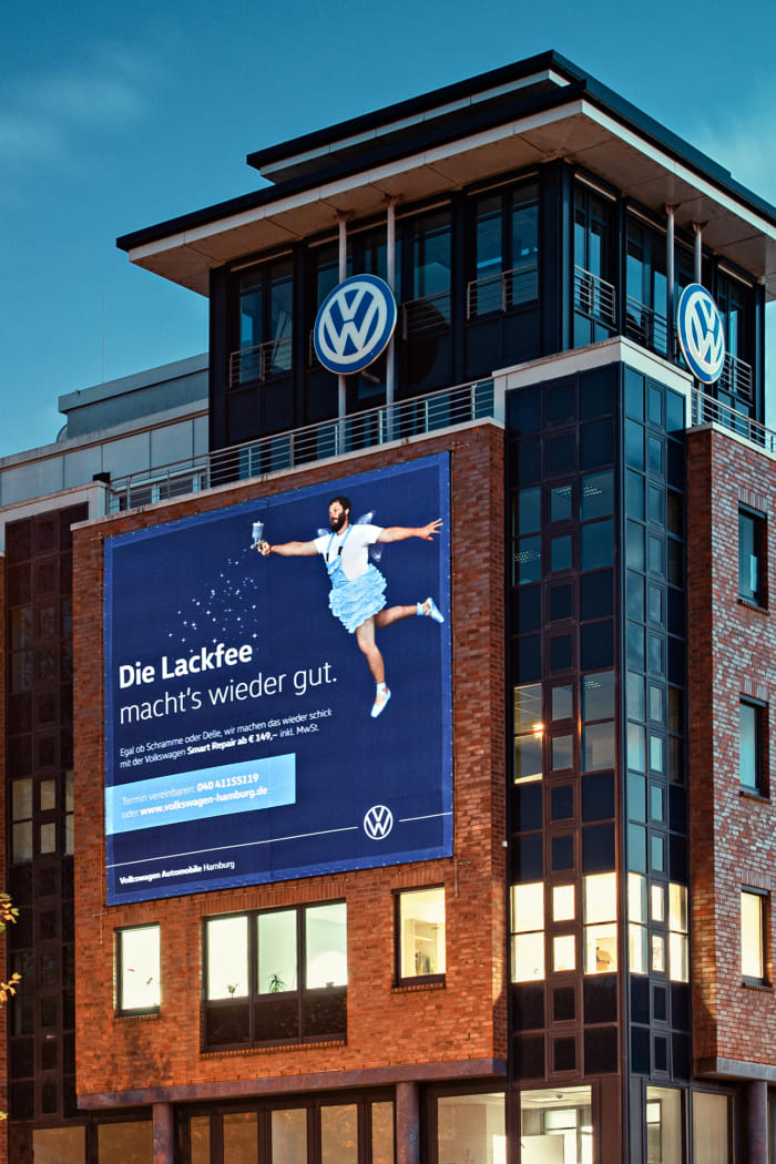 Die OOH-Fläche der Kampagne auf einem Gebäude in Hamburg zeigt ein Sujet mit blauem Hintergrund. Ein Mann in einem Tüllröckchen schwingt einen Zauberstab und fliegt optisch in das Sujet. Der Slogan der Kampagne ist ebenfalls abgedruckt.