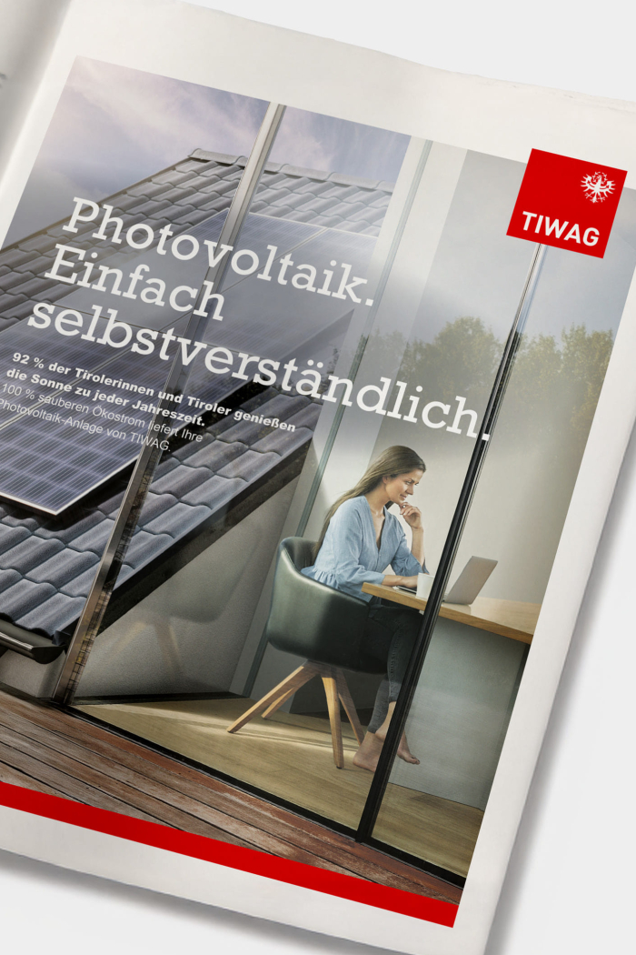 Eine ganzseitige Printanzeige zeigt eine Dame, die am Tisch sitzt und in ihren Laptop blickt. Im Hintergrund ist eine auf dem Dach montierte PV-Anlage sichtbar. Neben der Headline und dem Subtext der Kampagne ist das TIWAG-Logo abgebildet.