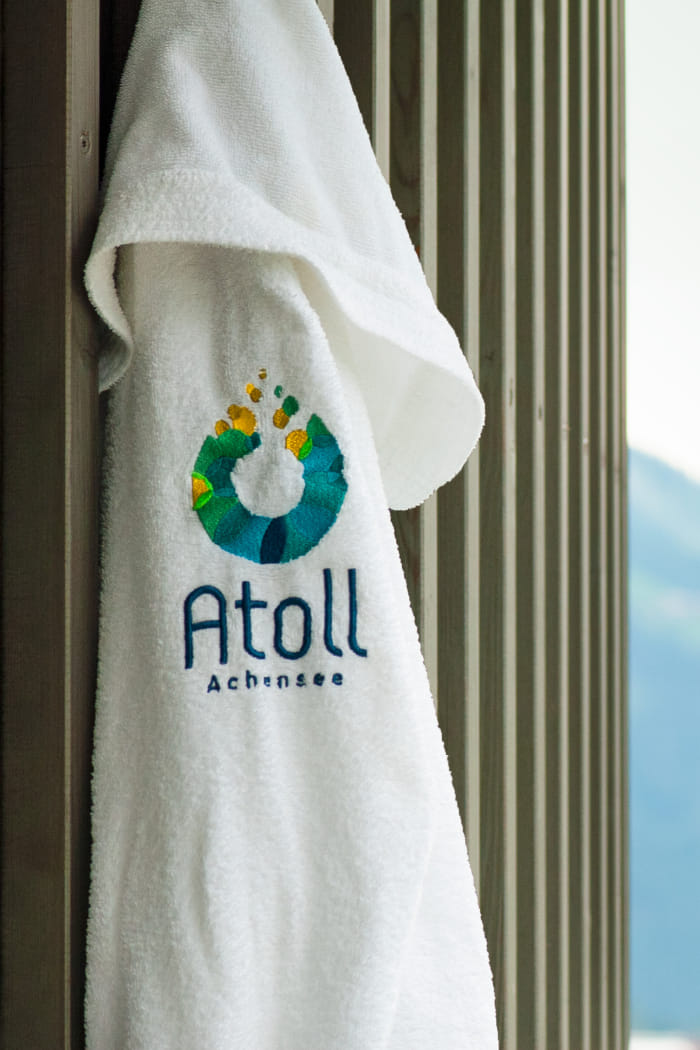 An einem Wandhaken auf einer Holzwand im Außenbereich hängt ein weißer Bademantel, auf dessen Rückseite das Atoll-Logo gestickt ist. Das Logo symbolisiert einen Wassertropfen, darunter steht der Schriftzug 'Atoll Achensee'.