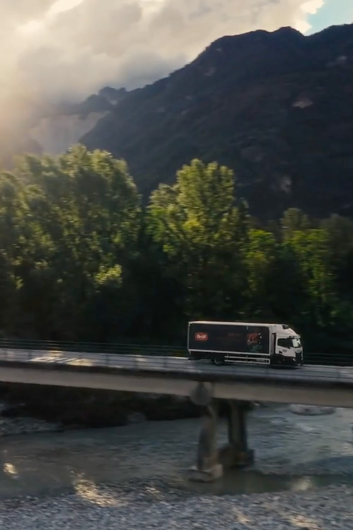 Mit dem Wedl Logo gebrandeter LKW auf einer Brücke, darunter fließt ein Fluss. Im Hintergrund sieht man grüne Bäume und Landschaft und die Sonne geht über die Berge auf.