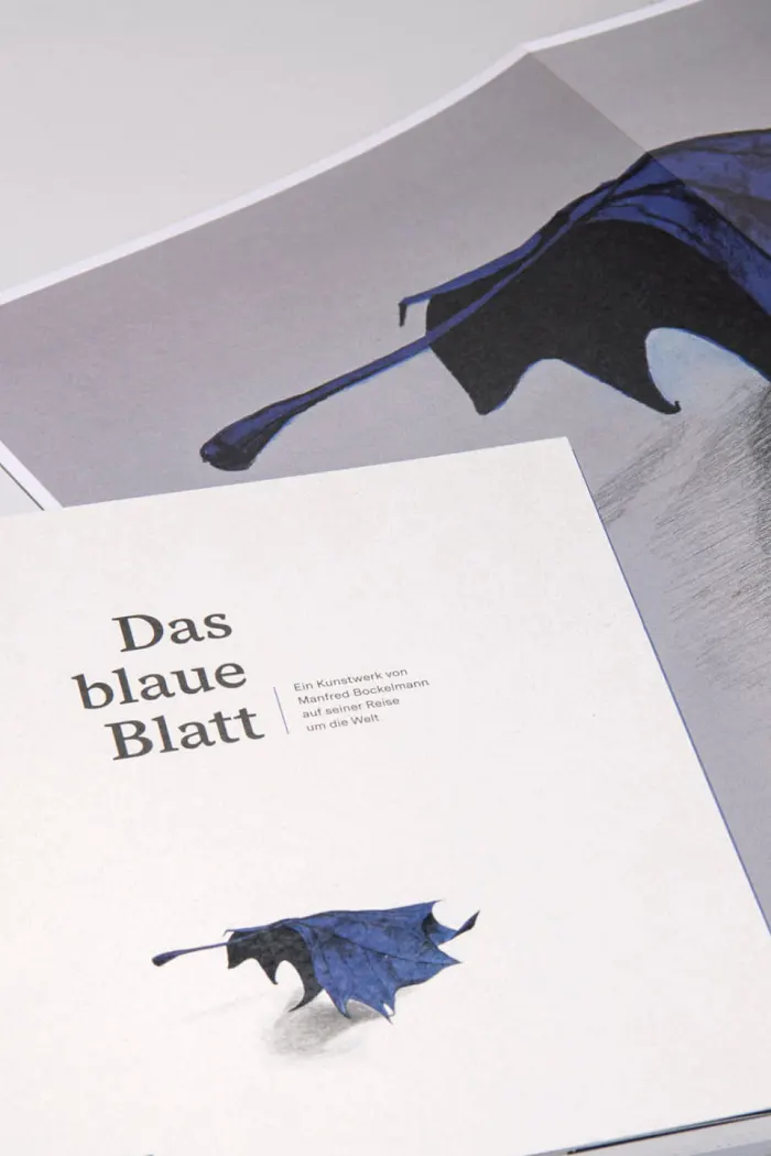 Auf weißem Untergrund liegen das Buch „Das blaue Blatt“ mit stilisiertem Titelmotiv und ein ausgebreitetes Poster mit einer detaillierten Zeichnung eines blauen Blattes