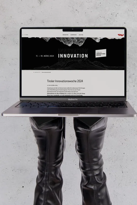 Vom unteren Rand des Bildes kommen schwarze Schuhe ins Bild, auf deren Sohle ein Laptop liegt. Auf dem Bildschirm des Laptops ist die Landingpage der Innovationswoche geöffnet.