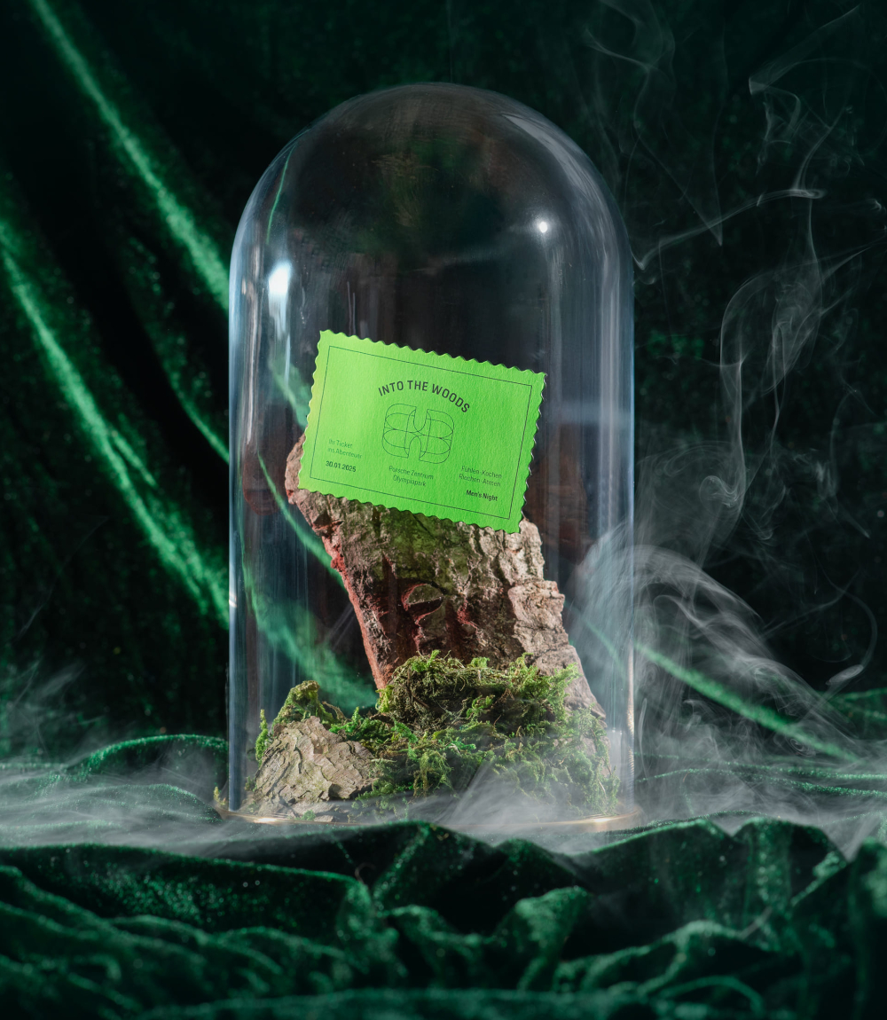 Unter einer Glaskuppel auf und vor dunkelgrünem Satin Stoff, befindet sich ein Baumstück und Moos. Weißer Rauch zieht vorbei und auf der Glaskuppel klebt ein grünes Ticket mit der Aufschrift INTO THE WOODS. 