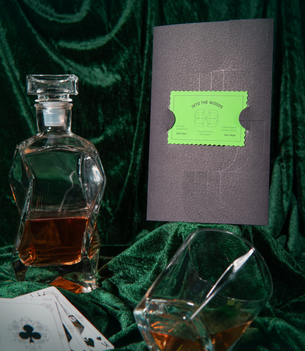 Auf und vor dunkelgrünem Satinstoff drapiert sind rechts eine schwarze Mappe in Lederoptik mit einem grünen Into the Woods Ticket mittig. Links eine halb gefüllte Whisky Flasche und vor der Mappe liegend ein Whiskyglas und drei aufgedeckte Spielkarten. 