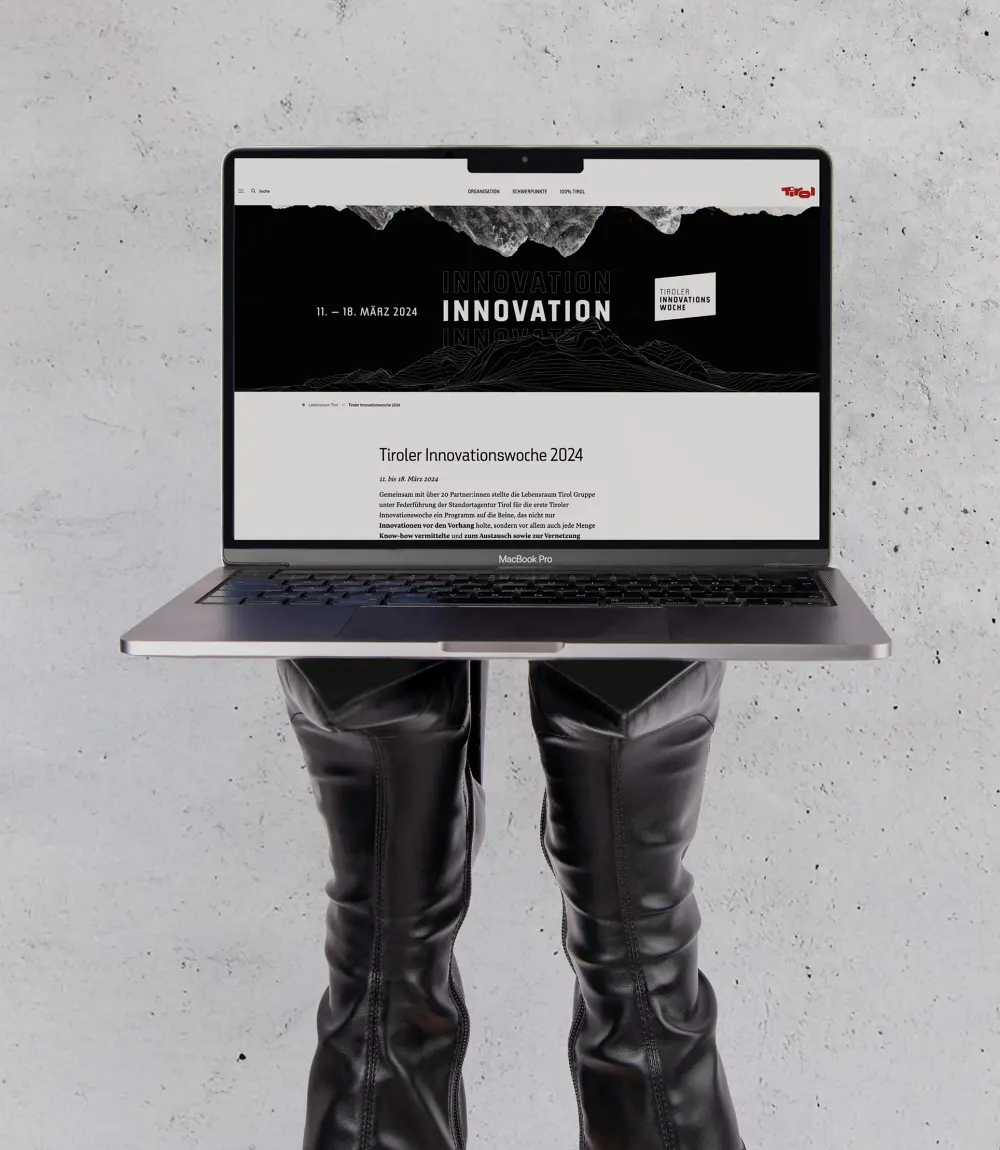 Geöffnetes Macbook das auf kopfüberstehenden Lederstiefeln balanciert wird zeigt die Website Tiroler Innovationswoche 2024.