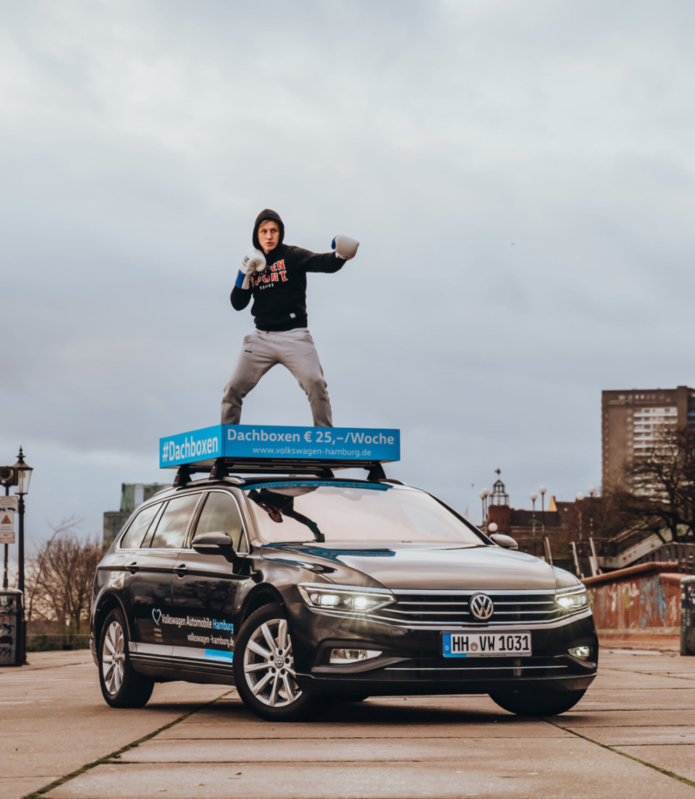 Junger Boxer in Kampfposition auf einer türkisblauen Dachbox mit der Aufschrift #Dachboxen € 25,-/Woche, die auf einem schwarzen VW Passat Variant montiert ist. Im Hintergrund ist das Hamburger Hafengelände bei bewölktem Himmel zu sehen.