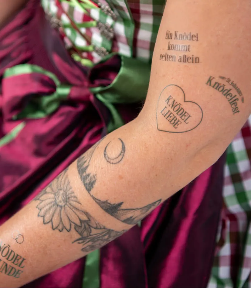 Eine Nahaufnahme eines Frauenarms zeigt eine Vielzahl an Klebetattoos, die alle in Bezug zum Knödelfest stehen.