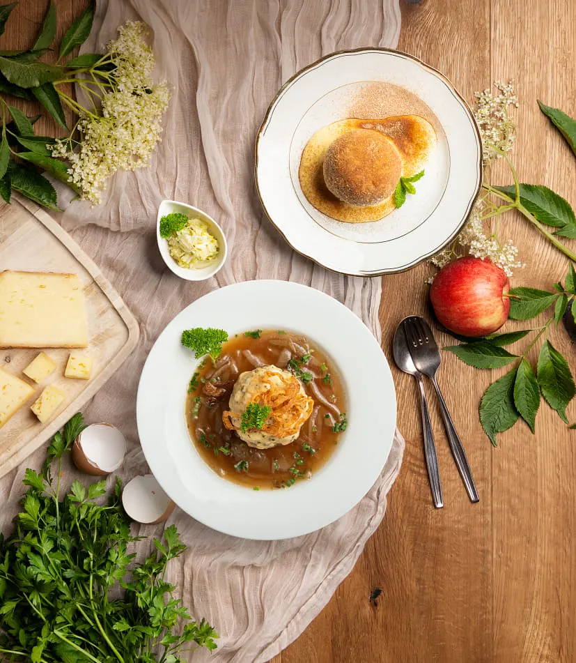 Auf einem dekorierten Holzisch sind zwei verschiedene Knödelsorten angerichtet – eine süße Variante und eine Suppeneinlage. Frisches Obst und Gemüse schmücken die Tischplatte und ergänzen die Szenerie.