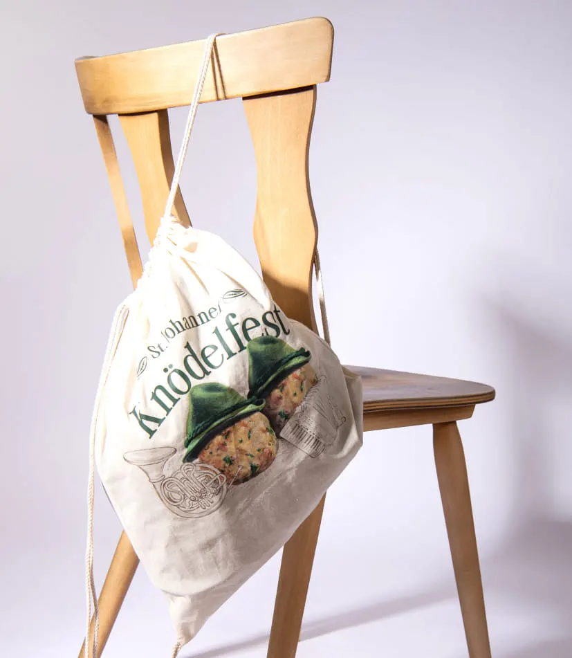 An der Rückenlehne eines Holzstuhls hängt ein Beutel aus Naturstoff. Auf dem Beutel ist das Logo des Knödelfests groß aufgedruckt – es zeigt zwei Knödel mit einem Trachtenhut sowie Illustrationen eines Blasinstruments (Horn) und einer Ziehharmonika.