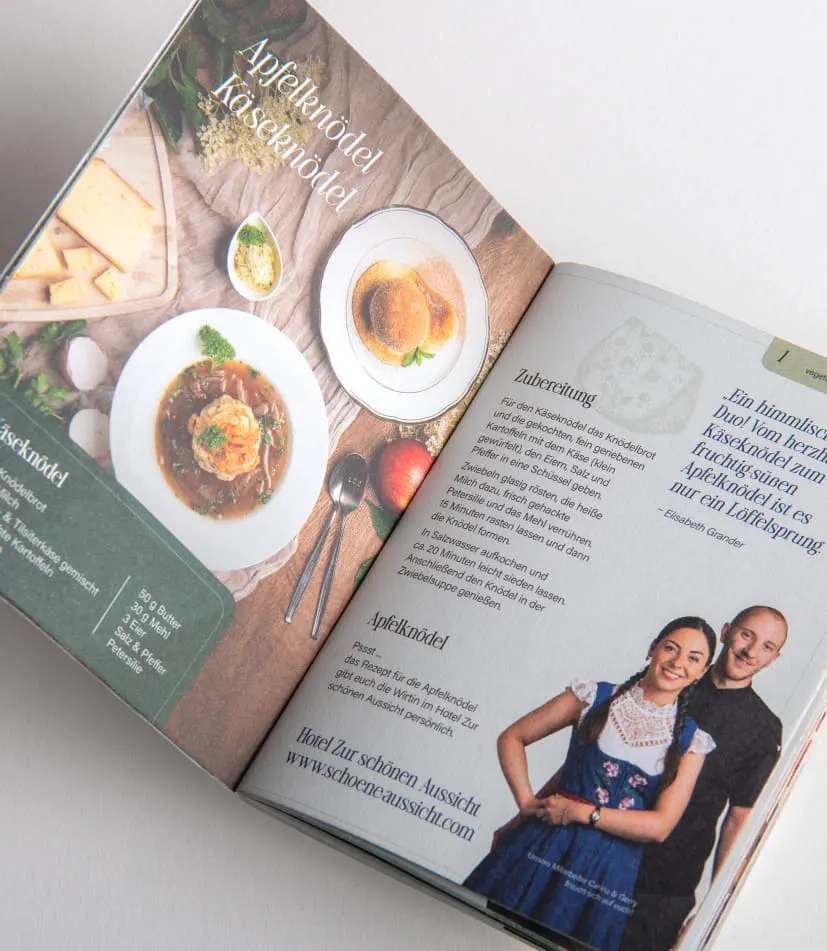 Auf weißer Oberfläche liegt das aufgeschlagene Rezeptbuch vom 42. Knödelfest. Die Doppelseite zeigt eine Fotografie der Knödel, rechts daneben ist das Rezept abgedruckt. Zusätzlich ist ein Portraitfoto der Personen zu sehen, die diesen Knödel beim Knödelfest zubereiten.