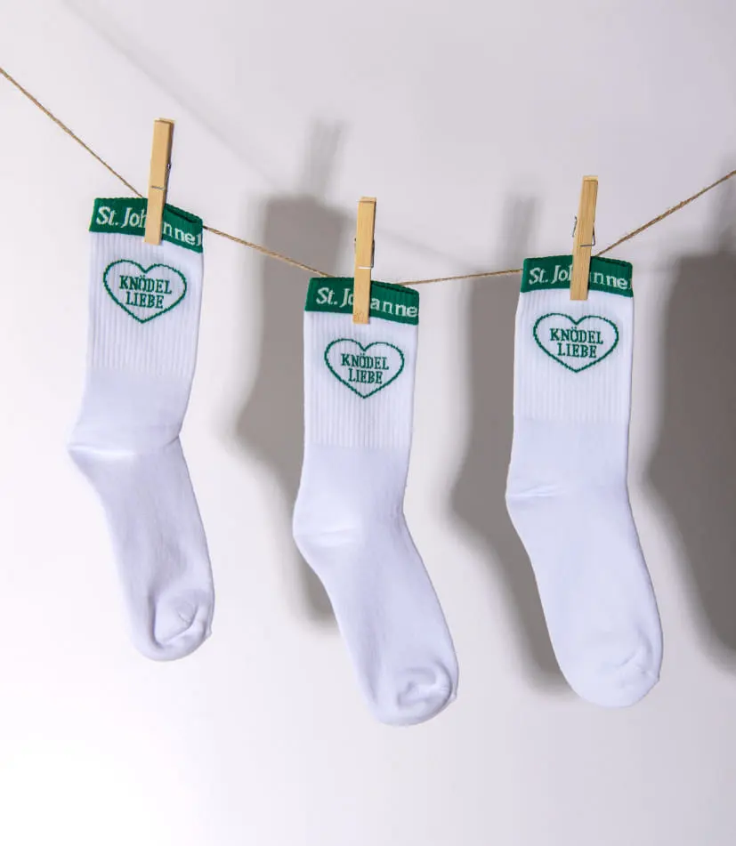An einer Wäscheleine hängen drei weiße Socken, befestigt mit Holzklammern. Der obere grüne Rand der Socken trägt die weiße Aufschrift 'St. Johanner Knödelfest'. Darunter ist ein grünes Herz eingearbeitet, in dem in Großbuchstaben 'Knödelliebe' steht.