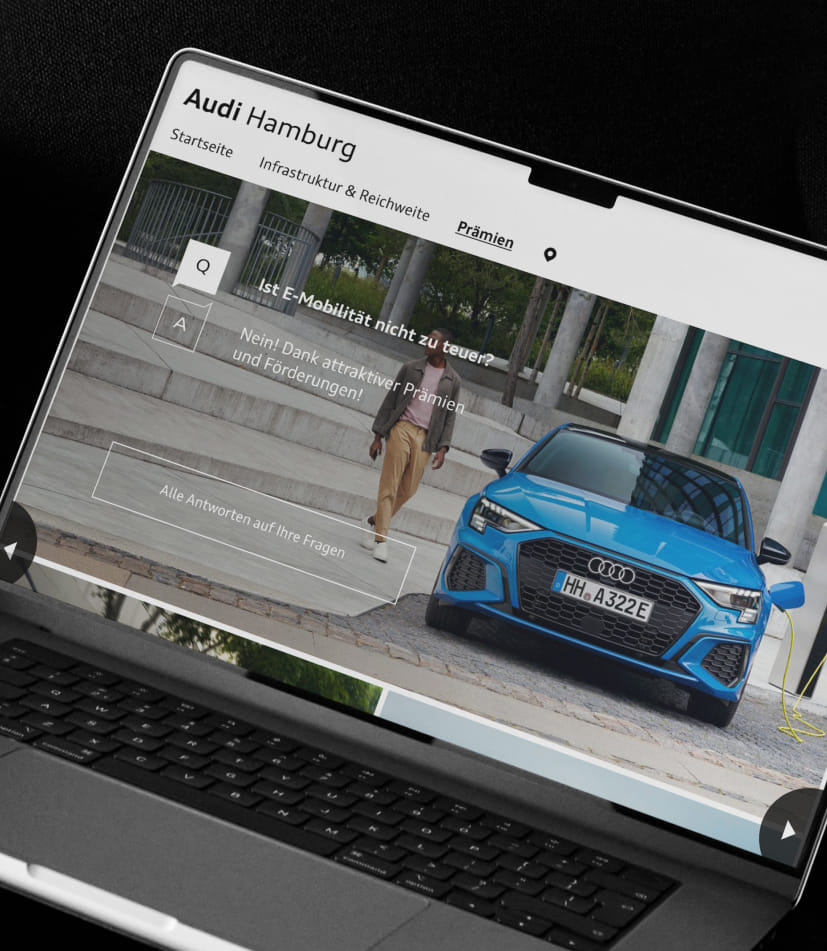 Auf einem Laptop ist die Audi Hamburg Webseite geöffnet mit der Frage: Ist E-Mobilität nicht zu teuer?, und der Antwort: Nein! Dank attraktiver Prämien und Förderungen!, neben einem blauen Elektroauto an der Ladestation.
