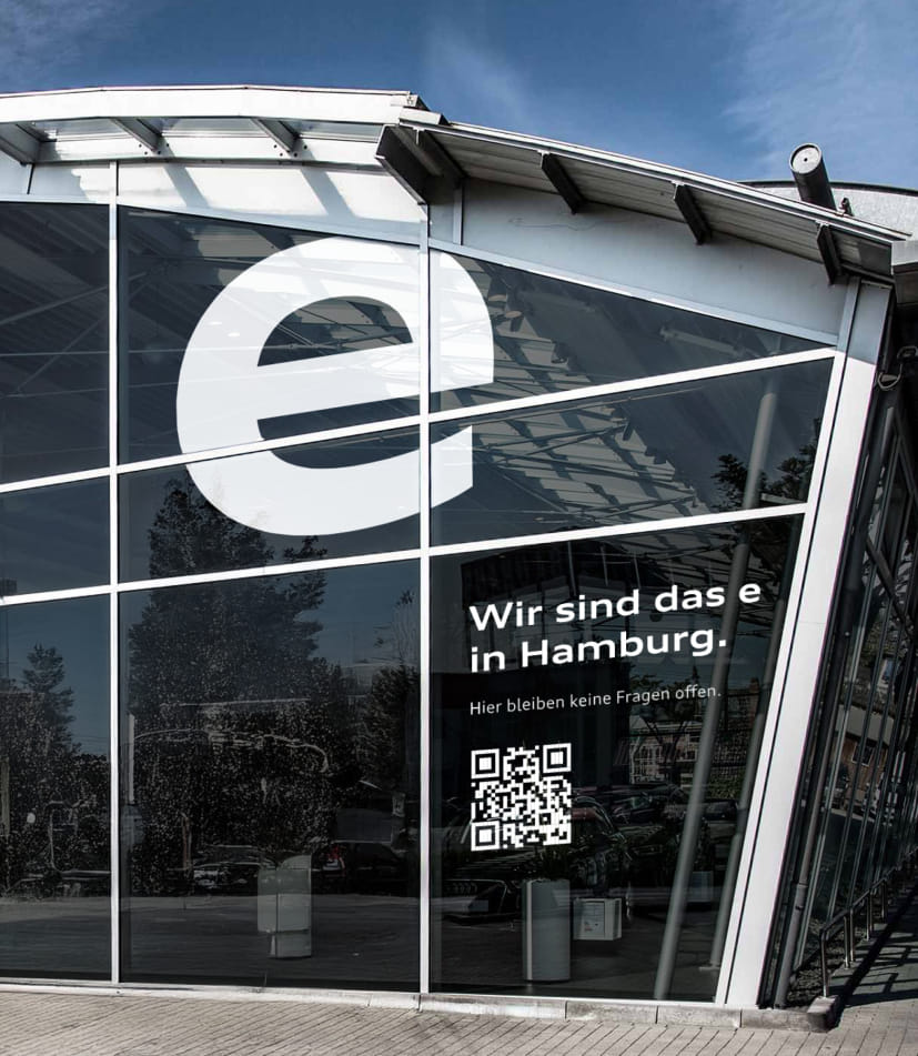 Die Glasfassade eines Audi-Autohauses in Hamburg mit großem weißen e-Logo und dem Kampagnenslogan: Wir sind das e in Hamburg sowie dem Zusatz: Hier bleiben keine Fragen offen, und einem QR-Code darunter.