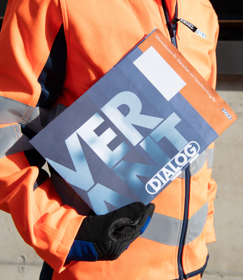Mitarbeiter in orangefarbener Arbeitskleidung mit Sicherheitsstreifen hält ein DAKA DIALOG Magazin. 