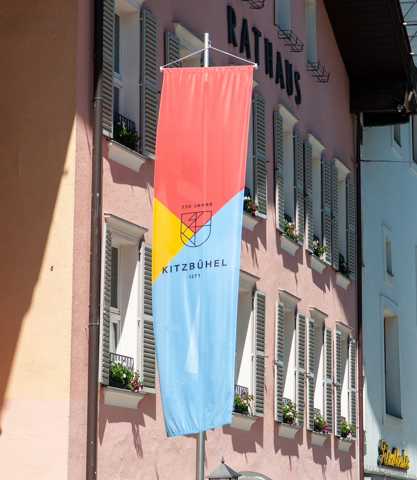 Vor dem Kitzbühler Rathaus mit roter Fassade und vielen Fenstern hängt eine Fahne im Layout des 750 jährigen Jubiläums. In der Mitte befindet sich ein stilisiertes Wappen mit grafischen Linien, darüber steht '750 Jahre', darunter 'KITZBÜHEL 1271'. Der Hintergrund ist in drei farbige Dreiecke unterteilt, oben rot, links gelb, unten blau.
