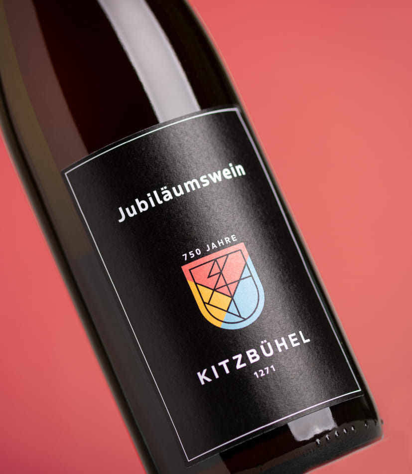 Nahaufnahme einer Weinflasche mit schwarzem Etikett vor rotem Hintergrund. Auf dem Etikett steht 'Jubiläumswein' sowie '750 Jahre Kitzbühel 1271'. In der Mitte befindet sich ein farbiges Wappen mit geometrischem Design in Gelb, Rot und Blau.