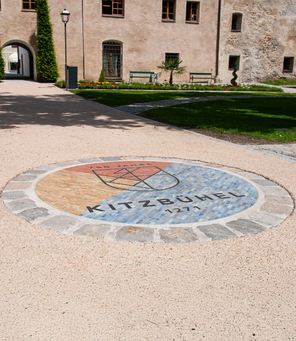 Rundes Bodenmosaik im Innenhof mit dem Jubiläumslogo „750 Jahre Kitzbühel 1271“. Das Mosaik zeigt ein stilisiertes Wappen auf farblich geteiltem Untergrund in Gelb, Rot und Blau. Im Hintergrund sind eine historische Gebäudefassade, grüne Rasenflächen und Parkbänke zu sehen.