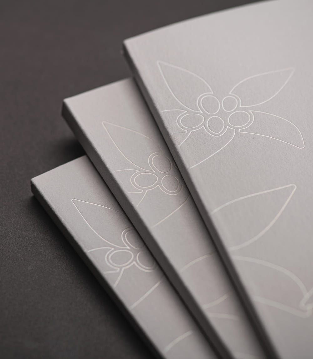 Nahaufnahme gestaffelter weißer Hotelbroschüren mit elegant geprägtem Blumenmuster. Die dezente Relieftechnik zeigt stilisierte Alpenblüten auf hochwertigem Papier.