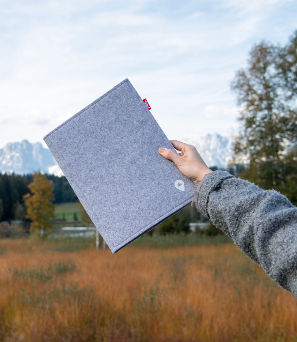 Gezeigt wird rechter Arm einer grau gekleideten Person auf einer Wiese stehend, die eine geschlossene Willkommen in Tirol Mappe hält.