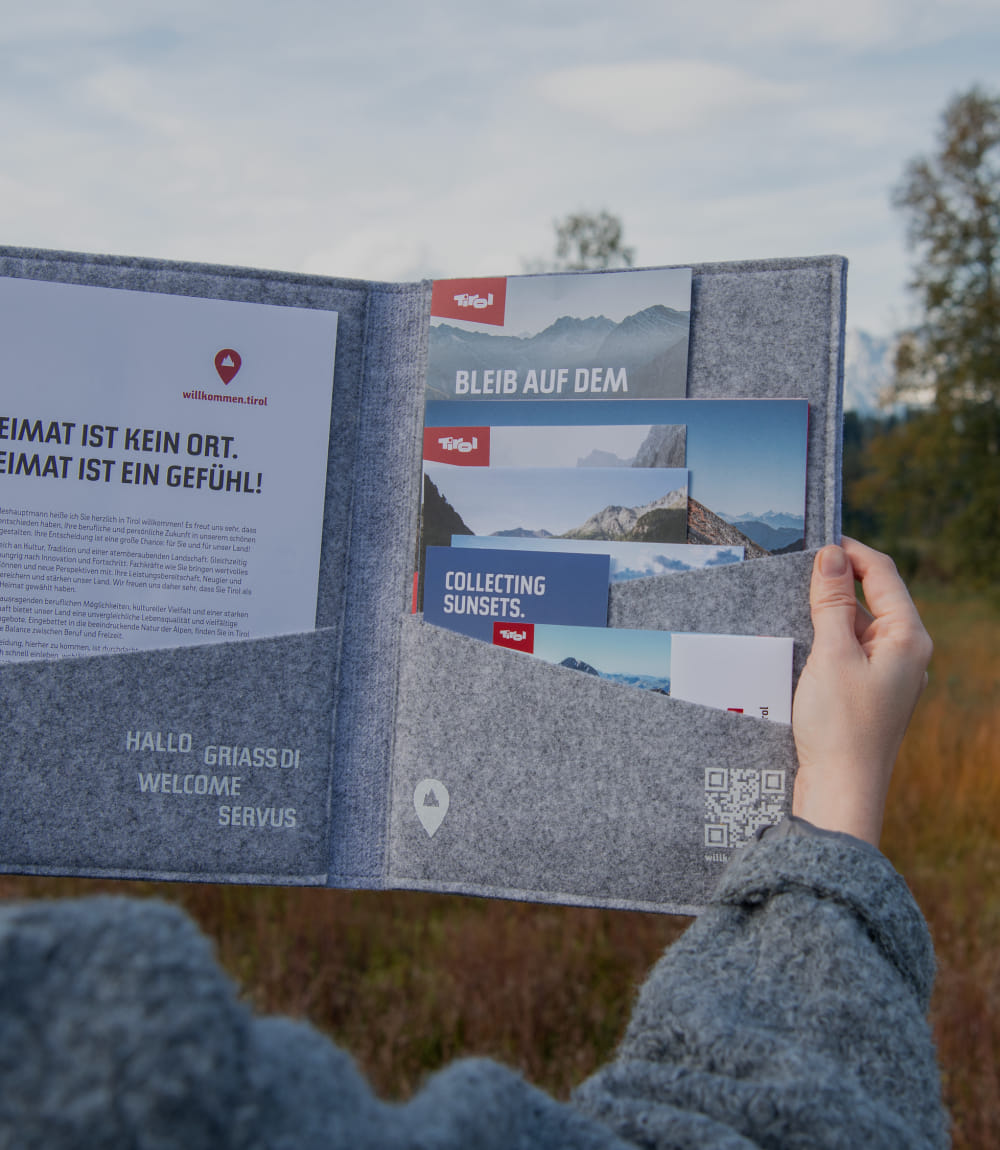 Gezeigt wird rechter Arm einer grau gekleideten Person auf einer Wiese stehend, die eine geöffnete Willkommen in Tirol Mappe hält.