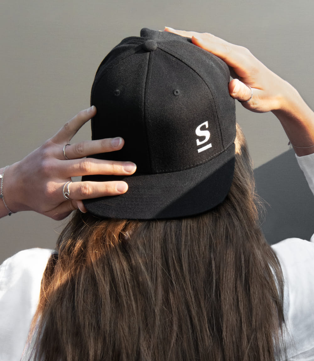 Junge Frau mit langen braunen Haaren und weißem T-Shirt von hinten zu sehen. Sie berührt ihre nach hinten gedrehte Baseball Cap mit beiden Händen auf der man das Sappl Logo sieht.