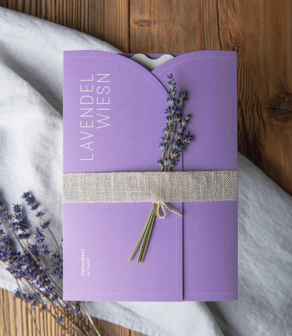Weißes Tuch auf Holzoberfläche. Darauf liegt ein lavendelfarbenes Kuvert verschlossen mit einem beigen Band. Ein Lavendelsträusschen ist in das Band geschoben und das Kuvert ist in weiß beschriftet mit LAVENDEL WIESN. Unter der linken Ecke des Kuverts liegt ein weiteres Lavendelsträusschen.