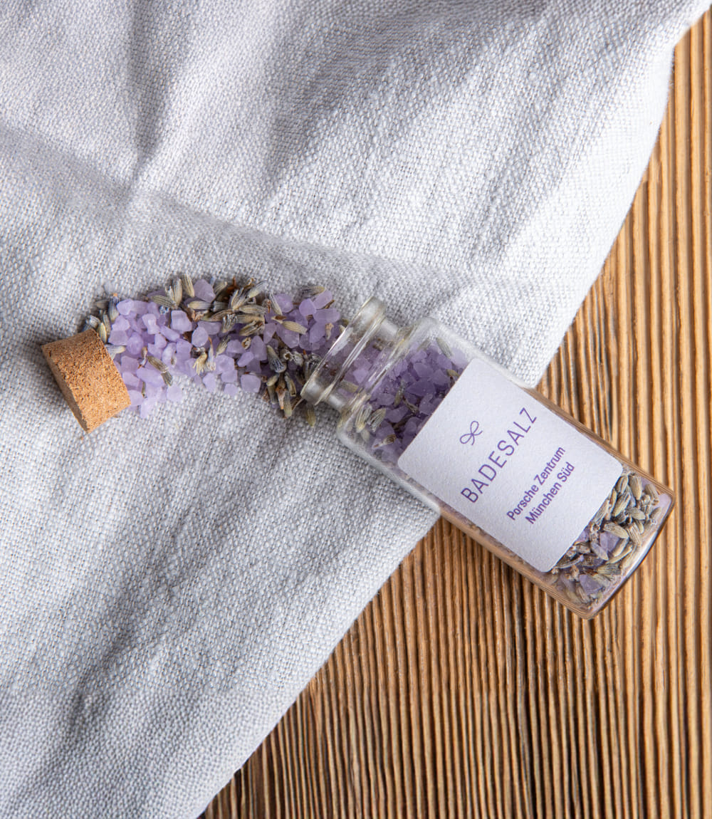 Auf einem weißen Tuch auf Holzoberfläche liegt ein geöffnetes Fläschchen mit Lavendel Badesalz das zum Teil ausgestreut wurde.