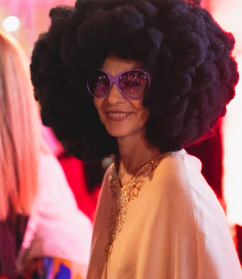 Frau mit großer Afro-Perücke und lila Sonnenbrille lächelt in die Kamera. 