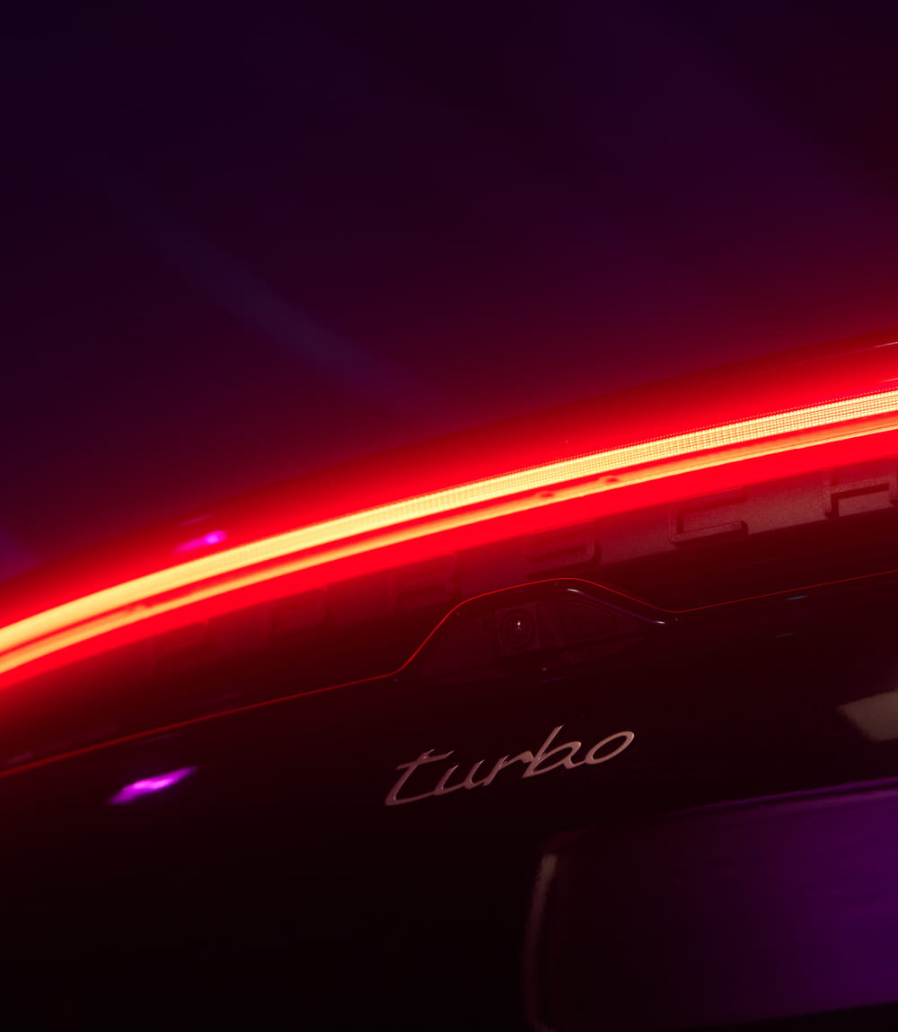 Die Heckansicht eines Porsche ist in sehr dunkler Umgebung zu sehen, wodurch der rote Lichtstreifen deutlich hervortritt. Darunter ist das Modell-Emblem 'Turbo' sichtbar.