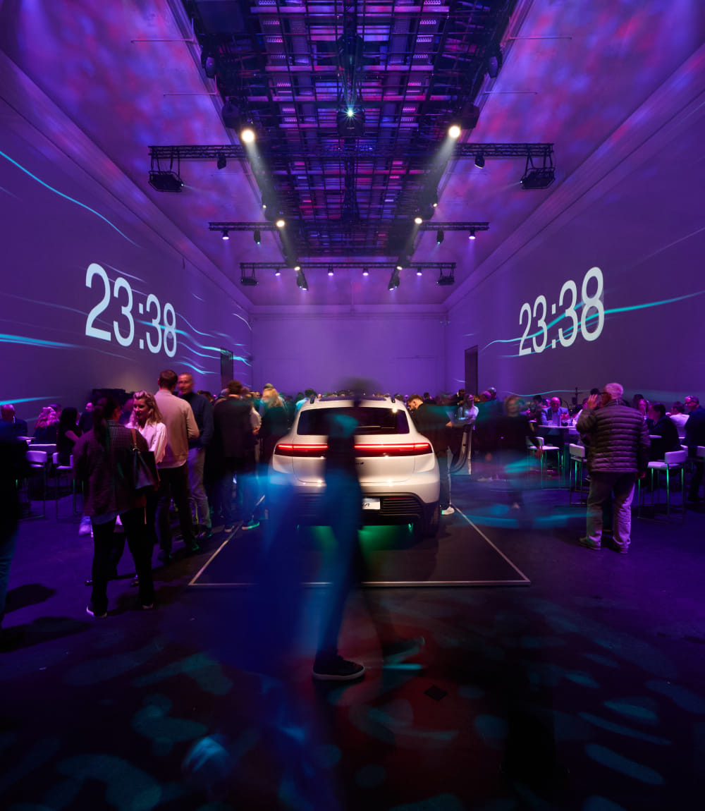 Der Veranstaltungsraum zeigt in der Mitte einen Porsche. Viele Personen sind leicht verschwommen im Raum wahrzunehmen. Die Projektion eines Timers zeigt eine digitale Anzeige mit ablaufenden Minuten und Sekunden, die in einer klaren, modernen Schriftart auf einer Wand projiziert wird.