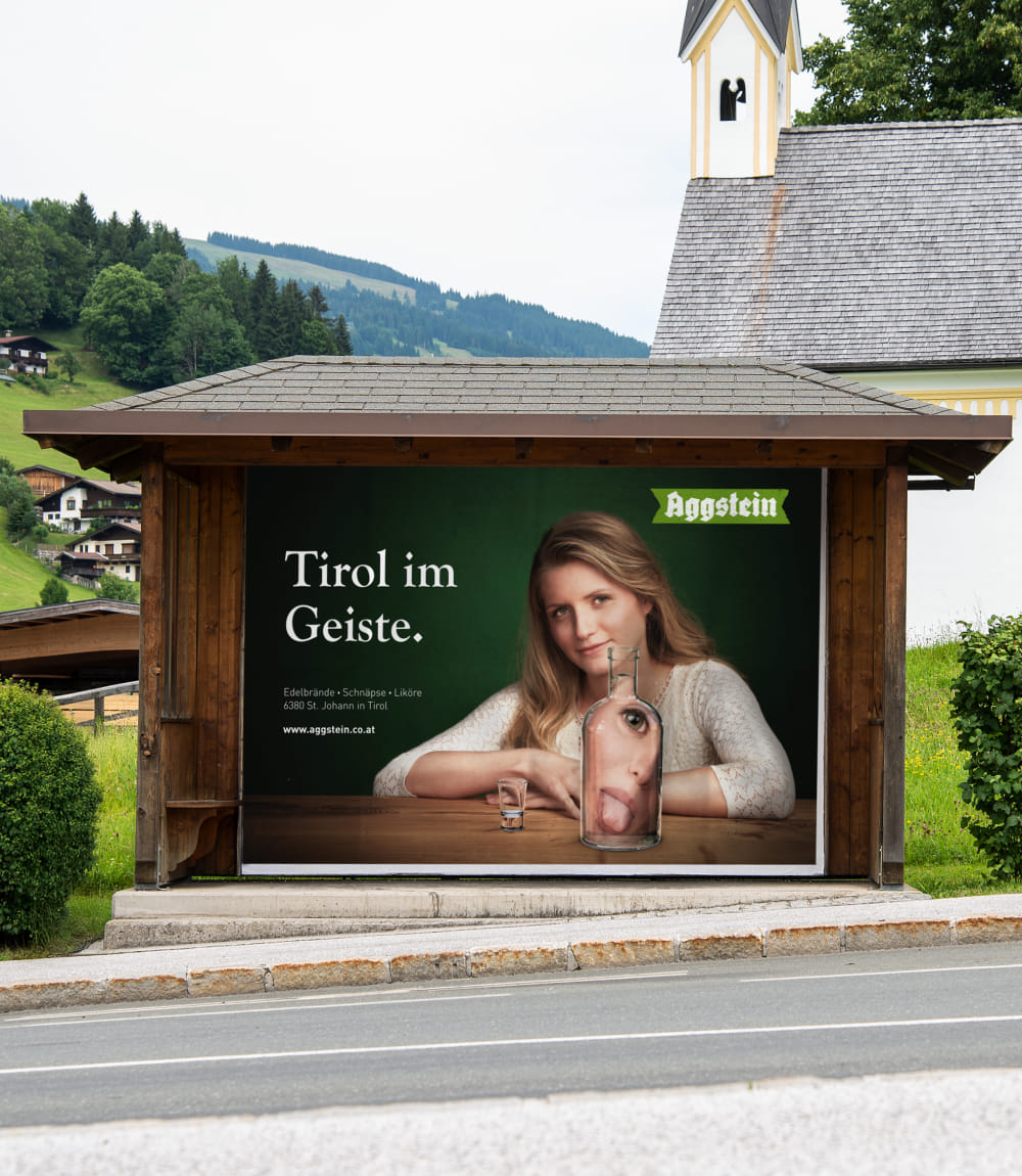 In einem Bushaltestellenhäuschen ist ein großformatiges Werbeplakat von Aggstein zu sehen. Darauf eine junge Frau mit blonden Haaren sitzt an einem Holztisch vor einer dunkelgrünen Wand. Ihr Gesicht wird in einer vor ihr stehenden Glasflasche mit rausgestreckter Zunge verzerrt dargestellt. Daneben steht noch ein passendes Schnapsglas.Links eine Schriftblock in weiß betitelt mit Tirol im Geiste.