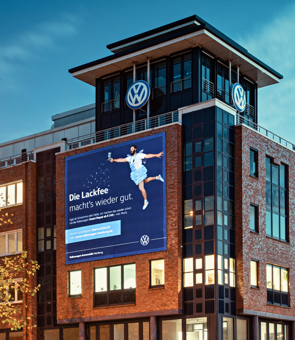 Quadratische Stoffwerbeanzeige zu Volkswagen Smart Repair auf VW Gebäude mit Backstein und Glasfassade. 