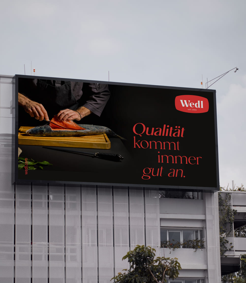 Gezeigt wird ein großes querformatiges Wedl Billboard in der oberen Hälfte einer weißen Hausfassade. Das Billboard zeigt die Hände eines Kochs in schwarzer Kleidung der einen frischen Fisch auf einem Holzbrett liegend filettiert.