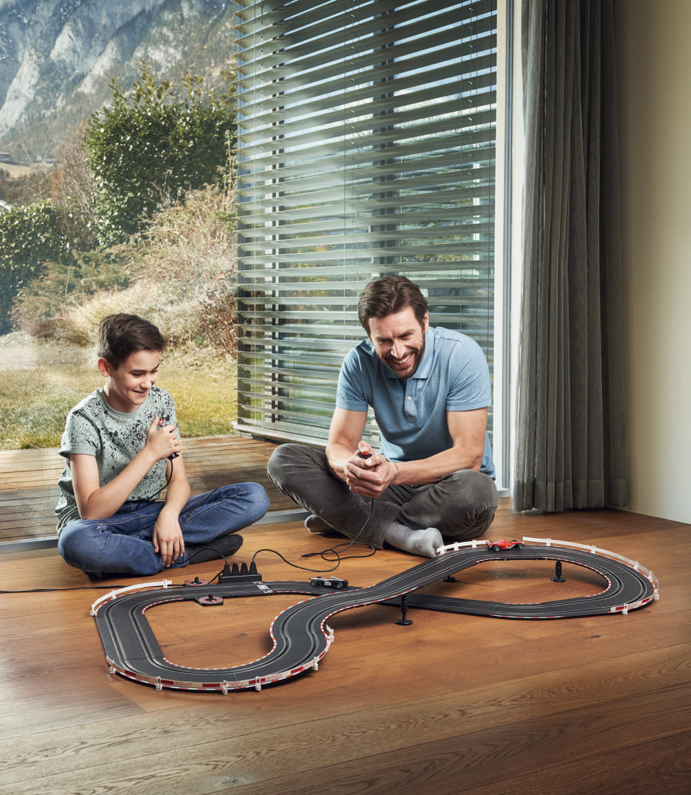 Vater und Sohn spielen gemeinsam mit einer Carrera-Rennbahn auf einem Holzboden vor einem großen Fenster mit Bergpanorama.