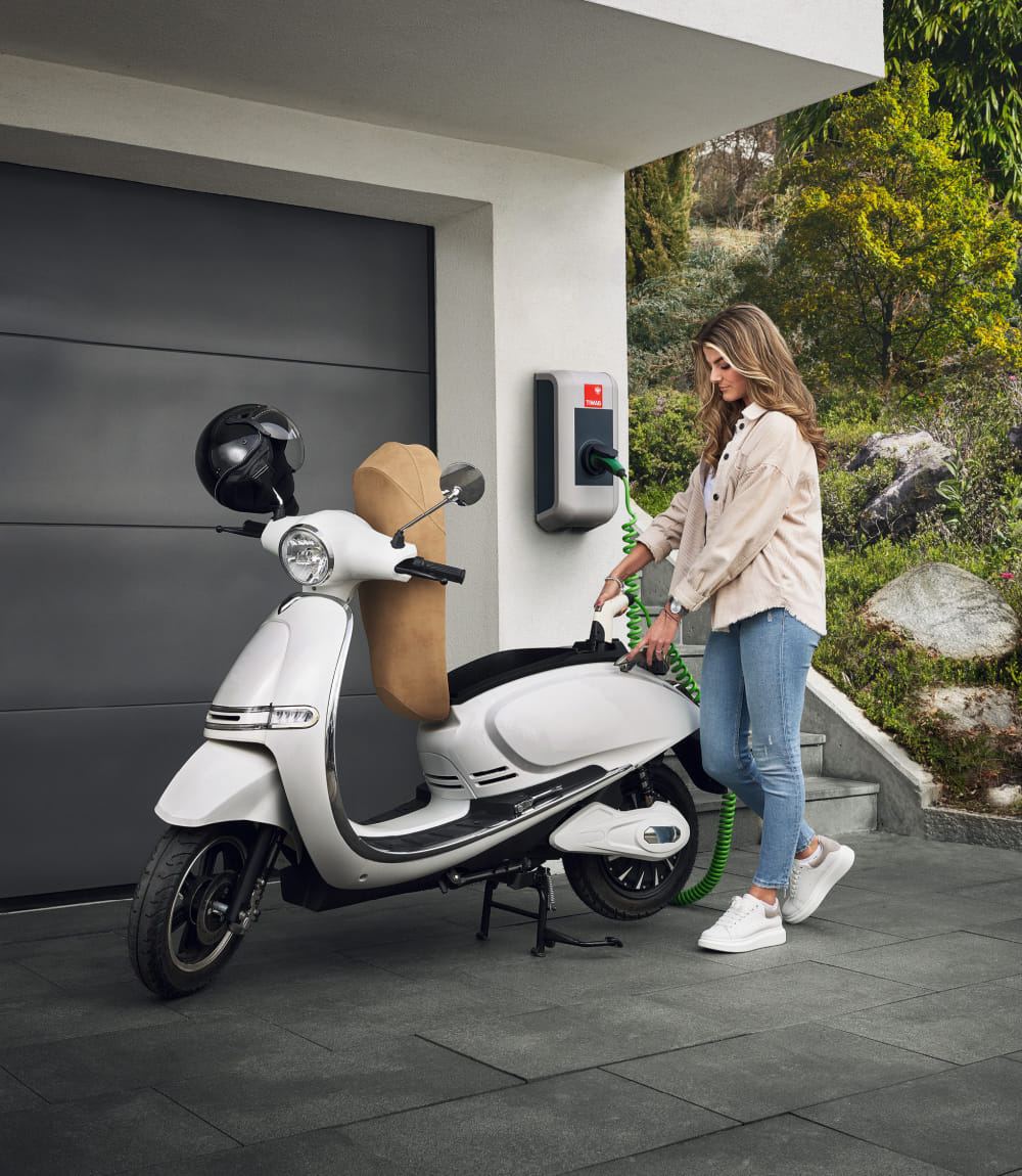 Junge Frau lädt einen weißen Elektroroller an einer TIWAG-Wandladestation vor einer modernen Garage mit grünem Naturgarten im Hintergrund.