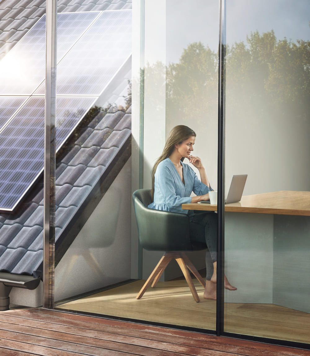 Frau arbeitet an Laptop in hellem Raum mit Glasfront, während draußen Photovoltaikmodule auf dem Dach sichtbar sind.