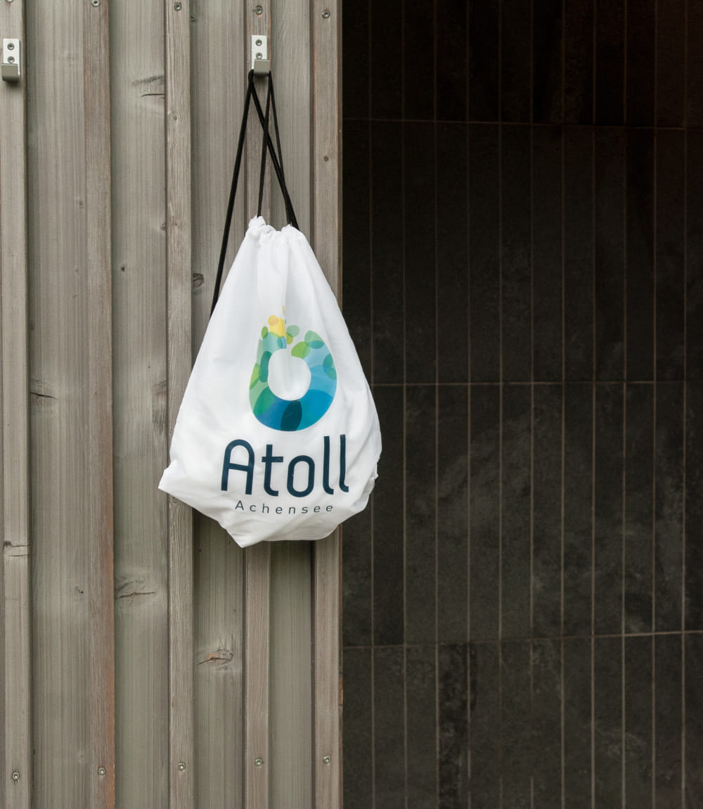Weißer Sportbeutel mit Atoll Achensee Logo an einer Holzwand hängend.
