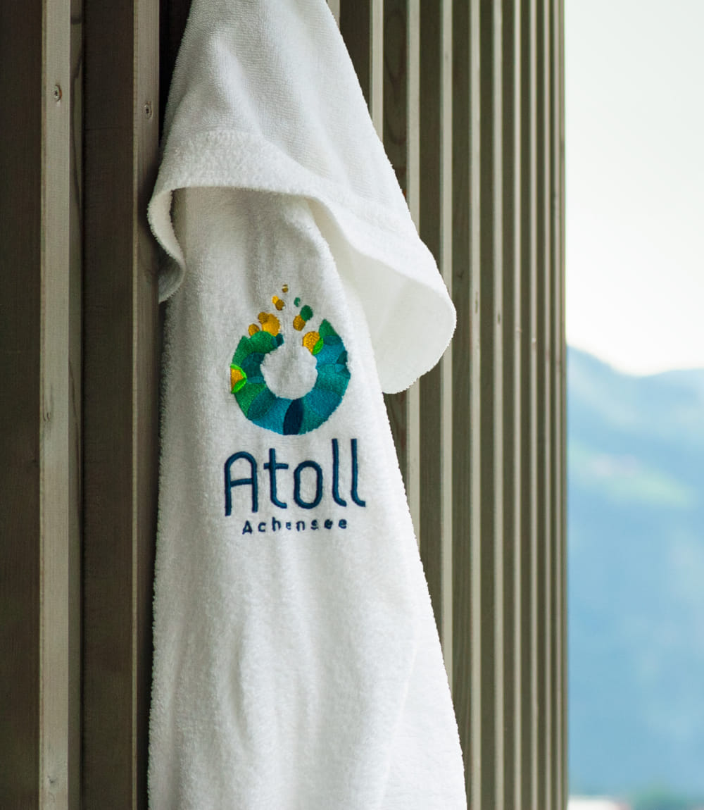 Weißes Handtuch mit Atoll Achensee Logo an einer Holzwand hängend. 