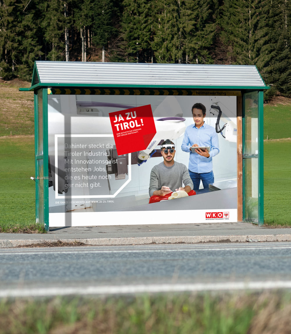 Bushaltestelle an einer Landstraße vor Tiroler Waldkulisse. Die Haltestelle zeigt eine Werbeanzeige der JA ZU TIROL! Kampagne mit rotem Logo. Auf dem Plakat sind zwei junge Männer in einem Innovationslabor zu sehen - einer mit VR-Brille, ein anderer mit Tablet. Der Text lautet: Dahinter steckt die Tiroler Industrie! Mit Innovationsgeist entstehen Jobs, die es heute noch gar nicht gibt.