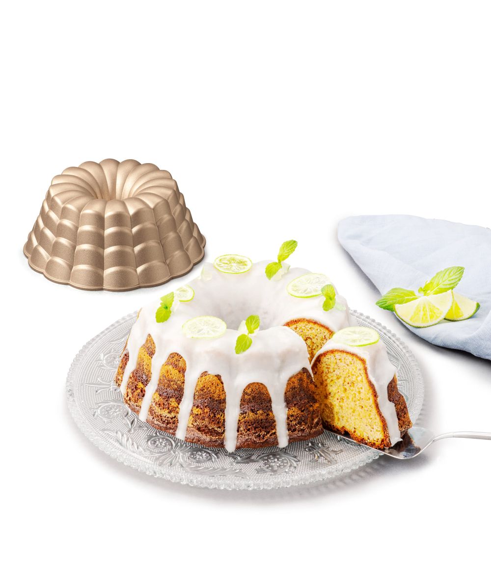 Goldene Gugelhupfform mit frisch gebackenem, glasiertem Kuchen auf Glasplatte. Dekoriert mit Limettenscheiben und Minze.