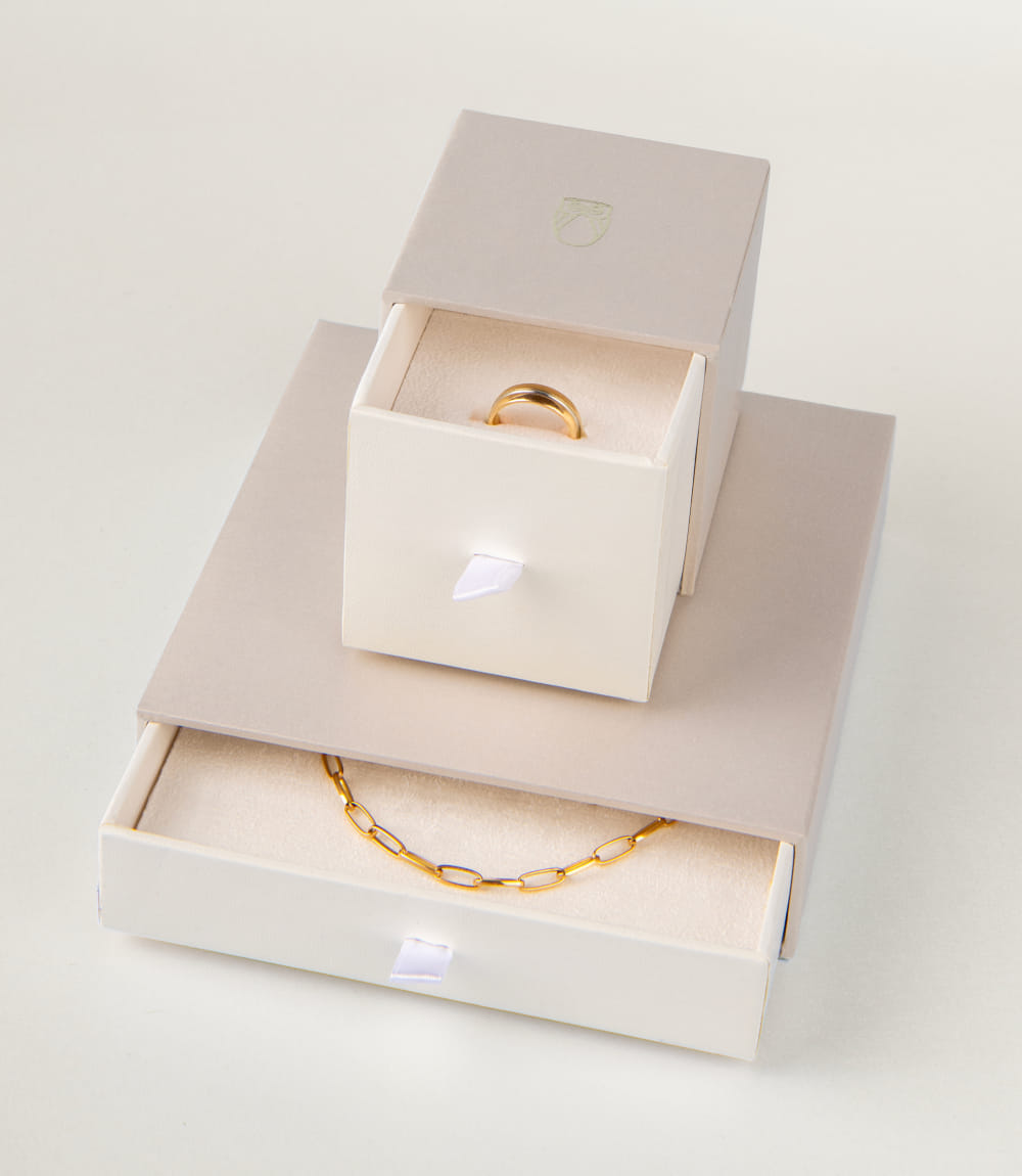 Zwei Schmuckverpackungen in Creme und Beige, eine mit goldener Halskette, die andere mit einem Ring.