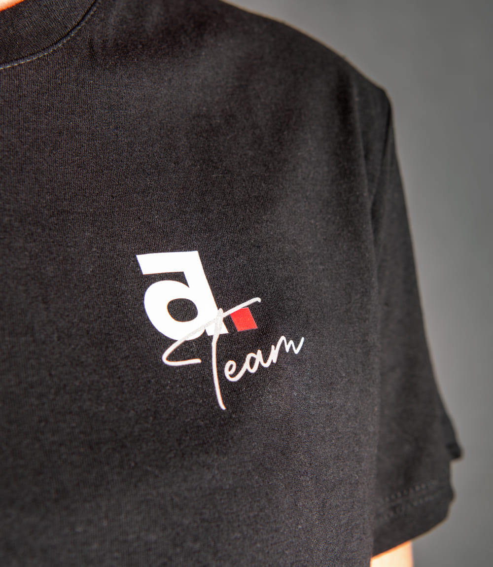 Vordere rechte Seite eines schwarzen T-Shirts vor grauem Hintergrund mit Schriftzug Team in weiß und kleinem weißen a mit viereckigem roten Punkt.