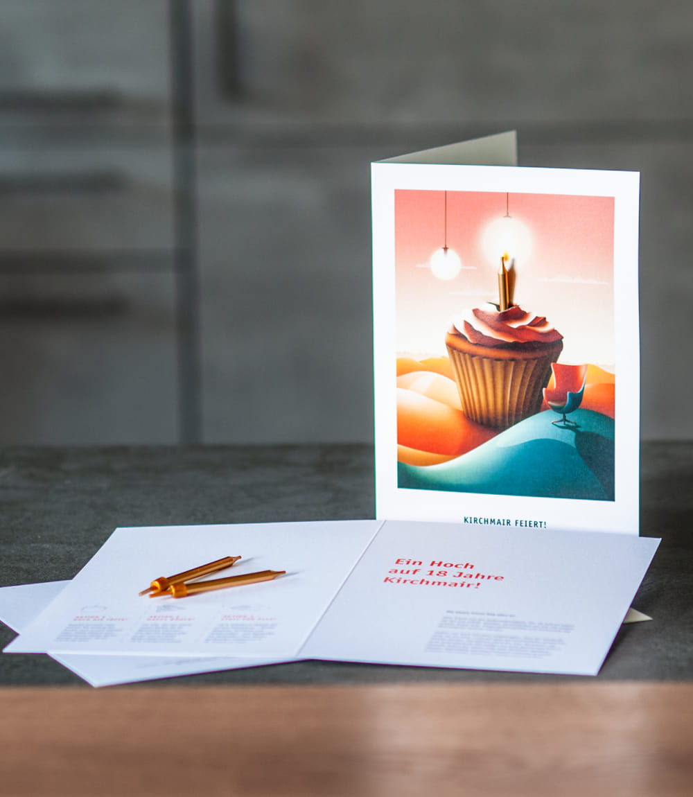 Auf grauer Stein- und Holzplatte steht eine aufgeschlagenen Grußkarte. Die Vorderseite zeigt eine Illustration mit einem großen Cupcake, einer Kerze und surrealen Elementen wie Hügeln, Leuchten und einem Designersessel. Daneben liegt eine geöffnete Grußkarte mit Text zum 18-jährigen Bestehen von Kirchmair und 2 goldenen Kerzen.