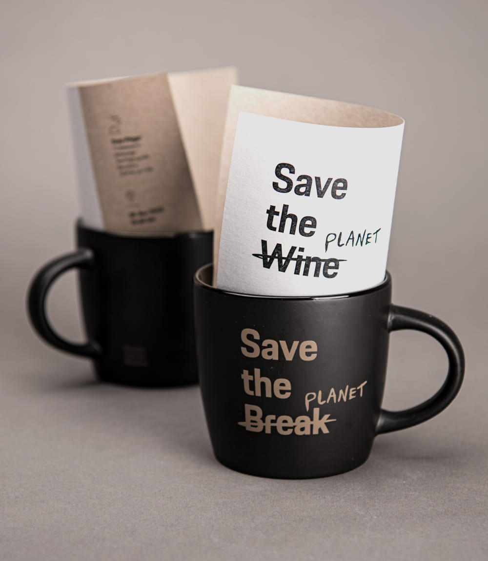 Zwei schwarze Tassen mit goldener Aufschrift stehen auf einem grauen Hintergrund. In der vorderen Tasse steckt ein gefaltetes Blatt mit dem Schriftzug Save the Wine – Wine durchgestrichen und ersetzt durch Planet, auf der Tasse selbst steht Save the Break – wiederum ersetzt durch Planet.