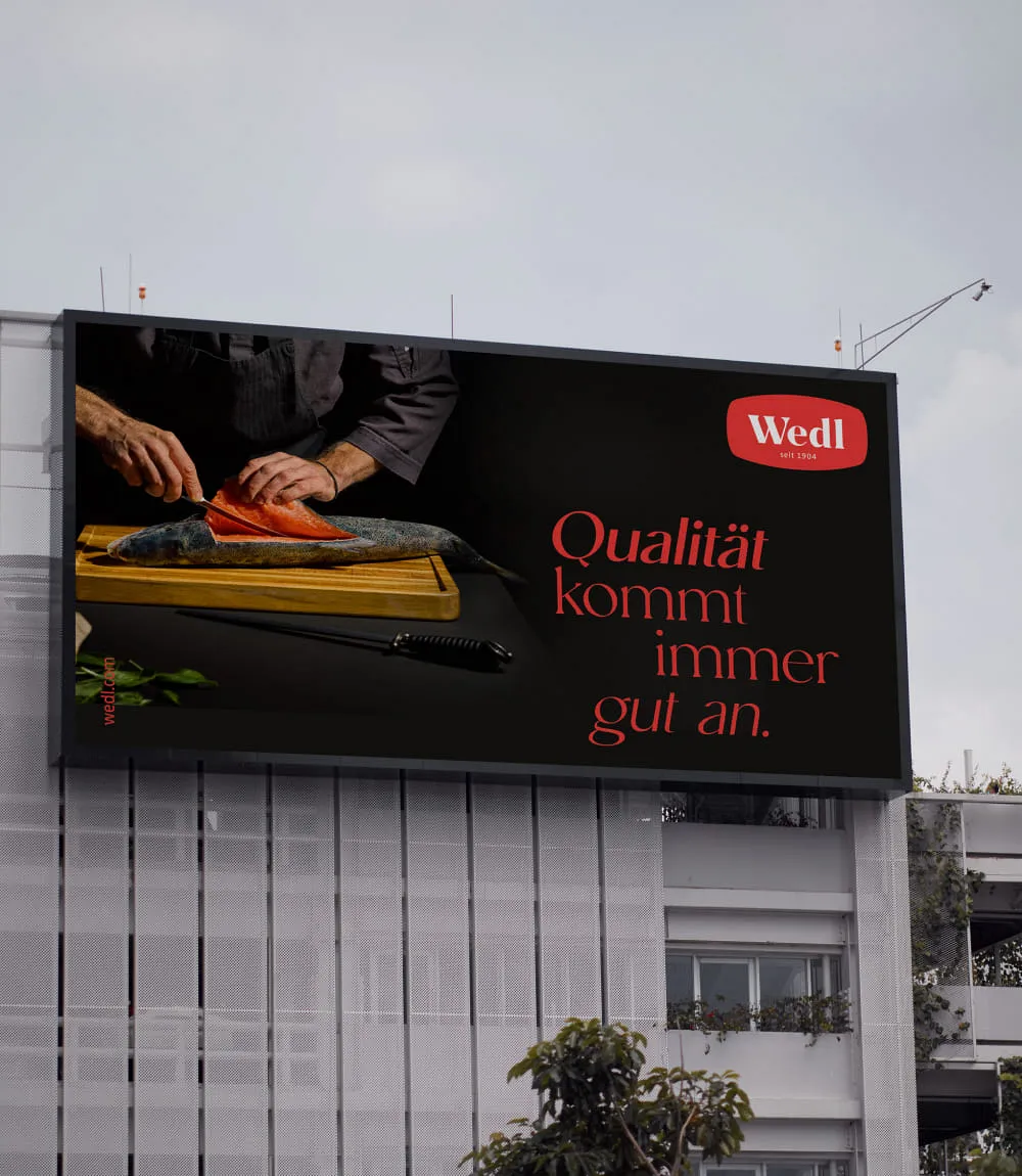 Gezeigt wird ein großes querformatiges Wedl Billboard in der oberen Hälfte einer weißen Hausfassade. Das Billboard zeigt die Hände eines Kochs in schwarzer Kleidung der einen frischen Fisch auf einem Holzbrett liegend filettiert.